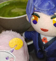 blurry photograph of plush doll of the character Leviathan, cat butler version, and sheep MC from Obey Me! One Master to Rule Them All. A teacup of matcha latte with a leaf foam art can be seen in the background.